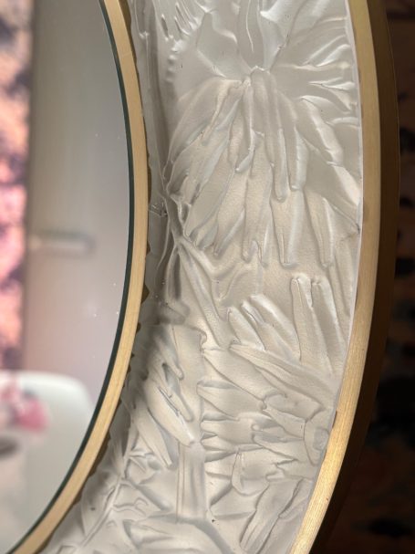 Miroir avec cadre décoratif en relief et doré, fond flou avec des fleurs.